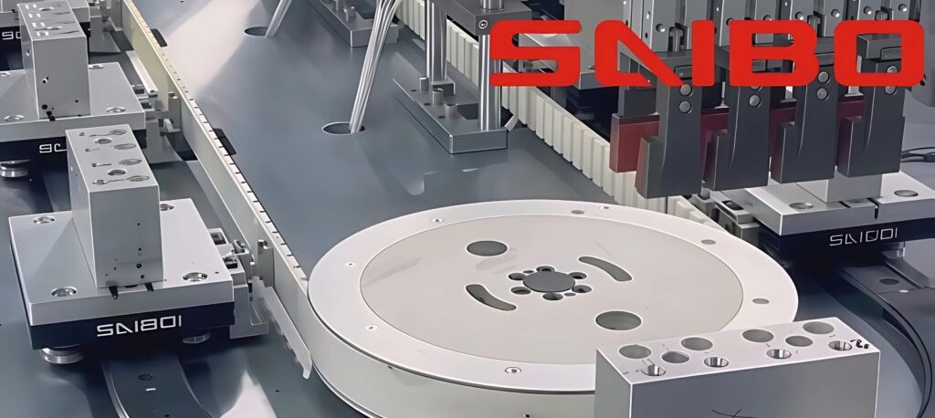 Pista oval de transferencia con correa dentada, platos circulares grises y carros negros sobre riel metálico, con logotipo SAIBO rojo en la plataforma.